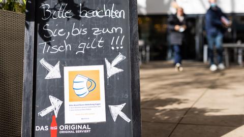 Ein Schild weist auf die Einhaltung der Maskenpflicht in einem Restaurant hin während im Hintergrund zwei Menschen zu sehen sind. (dpa)